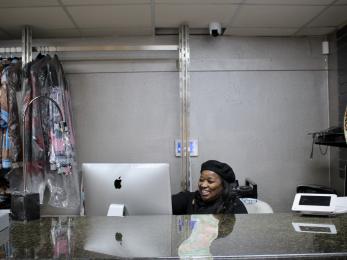 A black woman entrepreneur dressed in black sits behind her store counter with a smile