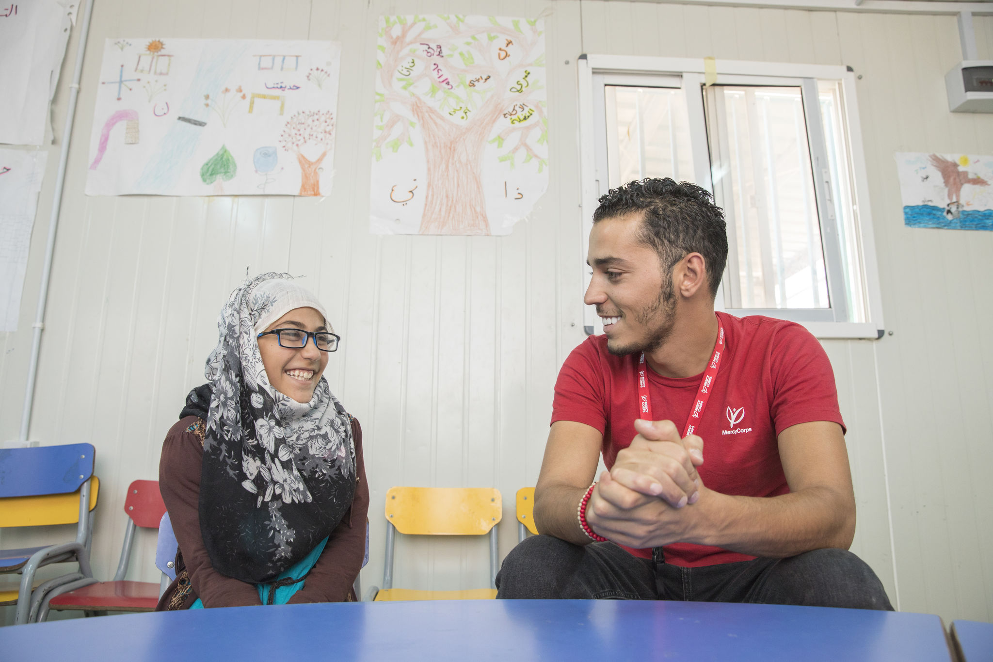 A Mercy Corps staff member sits at a table smiling with a young participant in Azraq, Jordan.