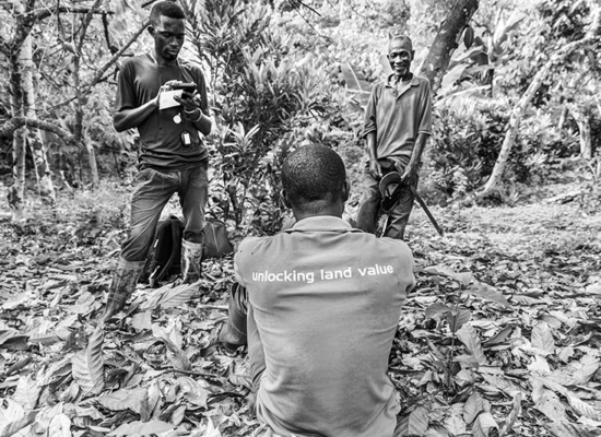 A person stands in a wooded area using a mobile device while another person sits on the ground and a third person stands nearby smiling.