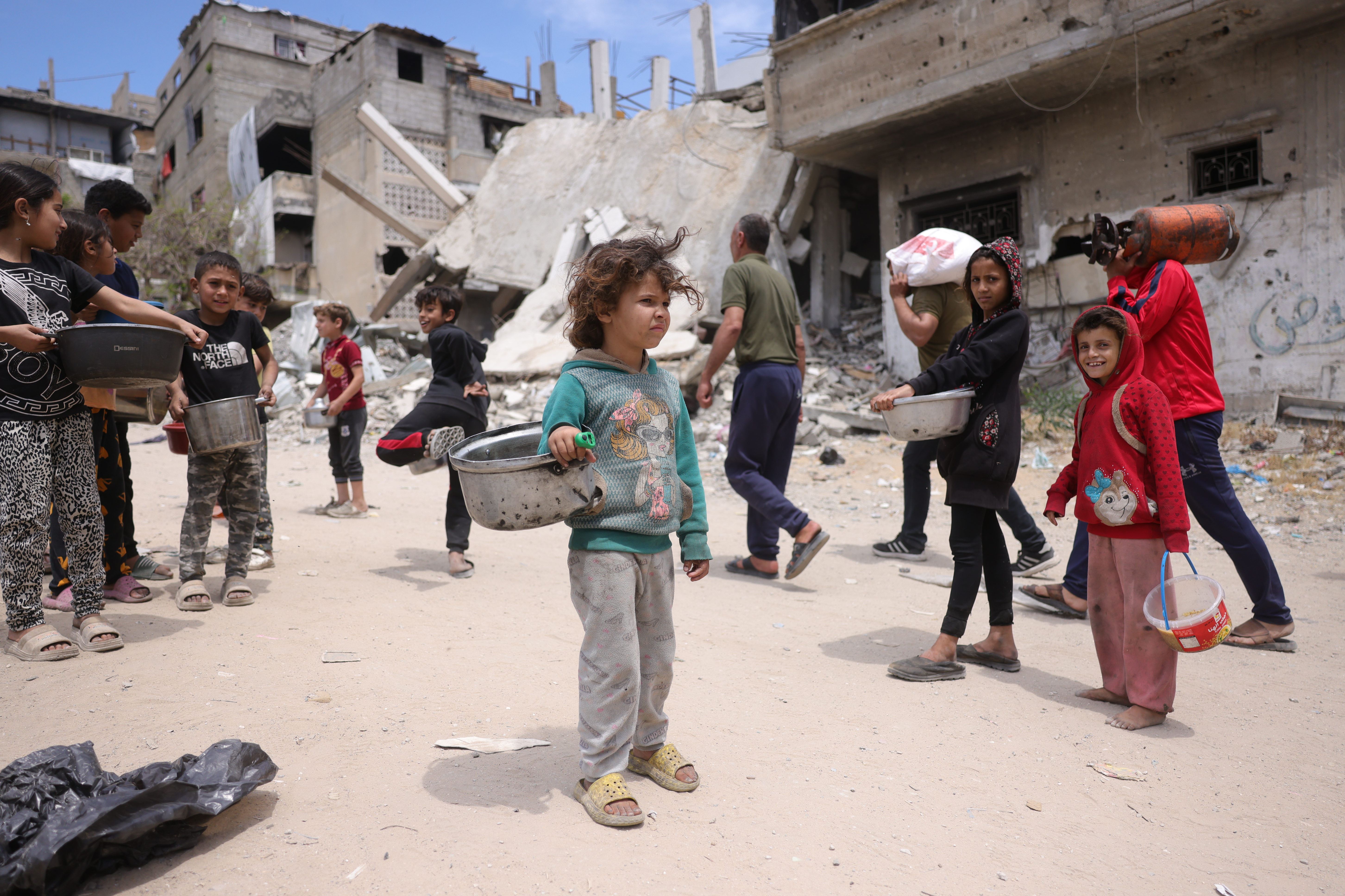 Palestinian children stand in a damaged public space with empty cooking pots.
