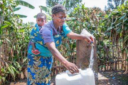 Mother and child collecting water from spigot in outdoor setting.