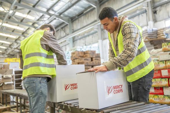 Two team members wearing safety vests package goods in boxes labeled with the mercy corps logo.