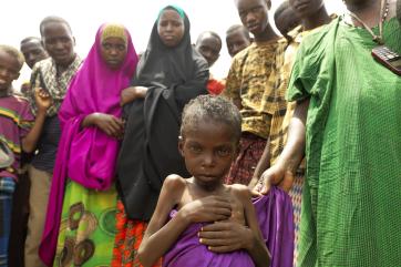 Young child wrapped in a purple cloth and looking seriously at the camera, with people standing behind them