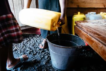 Hands pouring water from a yellow can into a metal bucket.