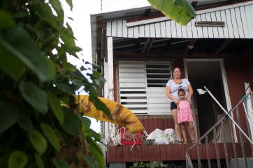 A woman rests her hands on the shoulders of a young girl. both stand on a front porch of a house.