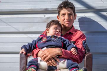 A young person sits in a chair while holding their sleeping younger sibling.