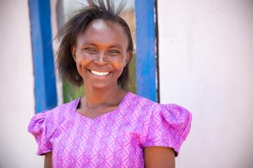 Woman smiling outdoors in front of a white wall with blue window frame.