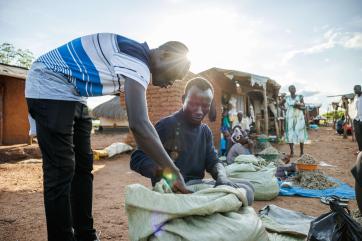 Moses aloro (striped shirt), a business mentor from village enterprise, interacts with his mentee, festo james (blue shirt), a south sudanese refugee who has set up a silver fish business in the local market.