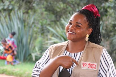 A mercy corps volunteer smiles at the camera and points to the organization's logo on her vest.