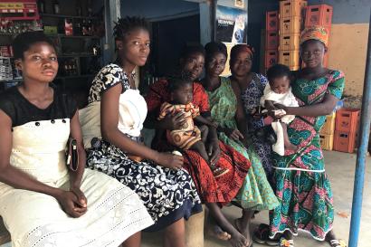 A group of people sit close together under an awning while some hold young children.