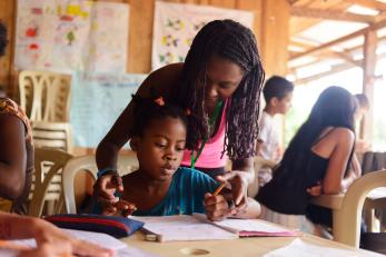 Carmen helping a child working in a notebook, seated at a table at school