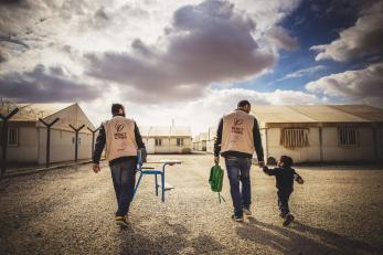 Two mercy corps team members and a small boy walking; one man is carrying a chair and the other man is carrying the boy's backpack and walking hand-in-hand with the boy.