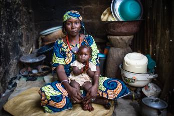 A woman sitting on the ground surrounded by pots and bowls, holding a baby on her lap. 