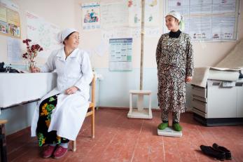 A woman stands on a scale in a medical facility. a medical professional is seated nearby taking notes. the two women are looking at each other and smiling.