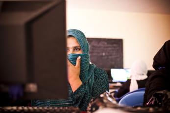 A girl covers her face beneath her eyes with her green headscarf as she works at a computer.