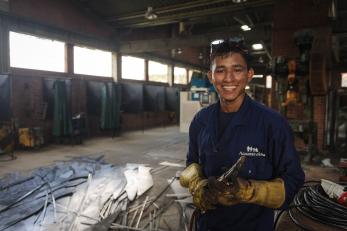 A young man in colombia pictured at the metal factory where he has undertaken an internship. he is wearing a blue work shirt and yellow gloves.