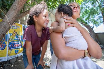 A woman holds her baby while her older daughter entertains the infant.