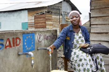A woman in drc next to a water tap labeled with usaid and mercy corps logos