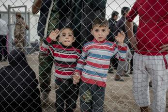 Two young boys on the other side of a chain-link fence, holding hands and wearing striped long-sleeved shirts