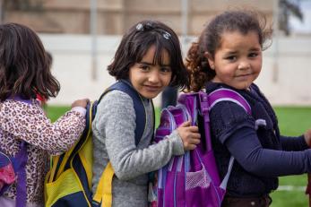 Young girls in jordan wearing backpacks, standing in line with their hands on the shoulders of the girl in front of them