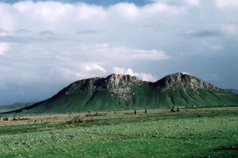 A green, flat plain leads to a rocky mountain formation against a cloudy sky