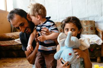 A father laughing with his young son while his daughter is nearby holding a blue stuffed elephant