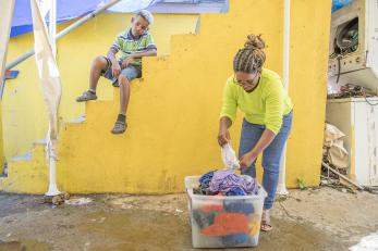 A boy sits on a yellow staircase with feet hanging over the stairs. his mother is sorting laundry below.