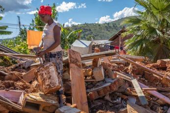 A woman holding a bucket stands among rubble under a blue sky dotted with clouds