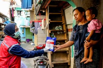 A mercy corps team member hands an aid package to an adult holding a child. 