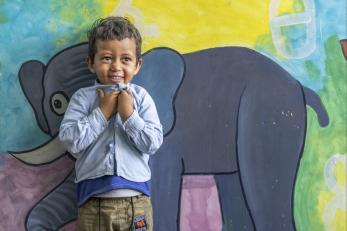 Ethiopian boy stands, smiling and clutching his shirt collar, in front of elephant mural.