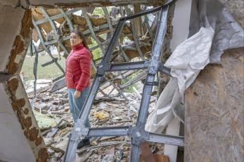 Ukrainian woman stands, staring at camera, through hole in wall of her home.
