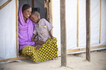 A woman wearing yellow and purple holds a toddler on her lap and sits in a doorway of a shelter