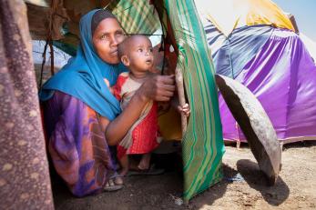 A parent and 9 month-old child look out from inside their tent at an idp camp.