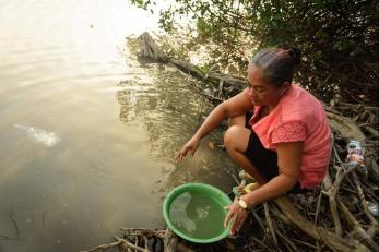 Oriana collecting water into a large green bowl from the river near her makeshift camp under the bridge.