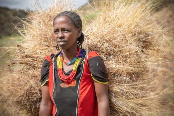 A woman wearing a colorful beaded necklace stands in front of a pile of straw in ethiopia
