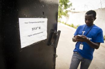 Image: a man in liberia outside the mercy corps office door. a sign reads: your temperature will be taken. 37.5 degrees c - higher will be turned away.