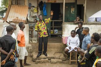 A young man addresses a group of other men in the community.