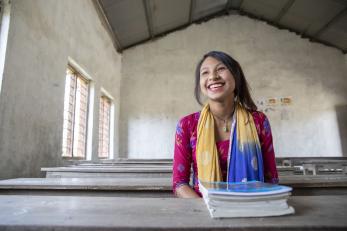 A teenager in nepal smiles widely while seated a table supporting a stack of books.