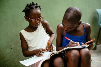 A young person holds a book for another while they write and use a device to complete school work in nigeria.
