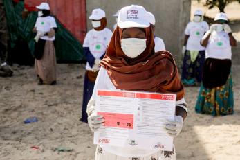 A person wearing a face mask and mercy corps hat holds an informational poster.