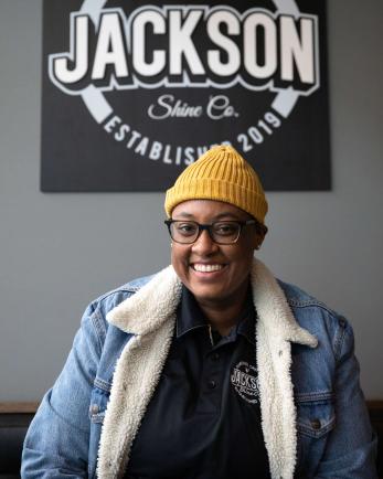 A small business owner smiles for the camera in front of a sign for their shoe shine business.