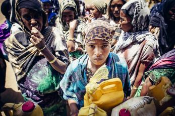 Women in scarves gathered around one woman, center, who is looking at the camera and holding a water container