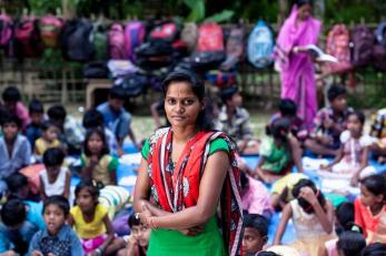 Woman standing with arms folded in front of children seated on floor. backpacks hanging in background.