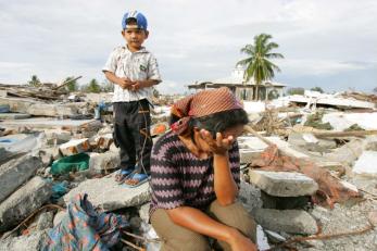 Small boy stands on rubble in background. In foreground, a seated woman wipes away tears.