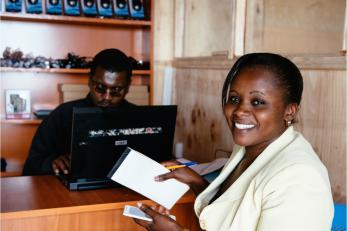 Lucy at a counter across from a man on a computer, smiling and holding a cell phone, notepad and pen.