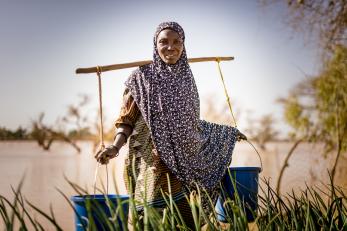 Woman in patterned scarf balancing two blue buckets across her shoulders using a stick