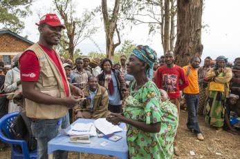 Mercy corps team member speaking to a woman from behind a blue table. the woman has a baby sleeping, swaddled on her back.