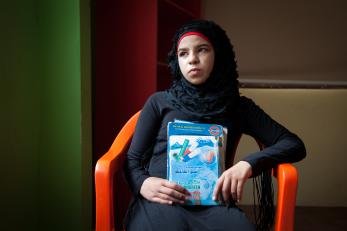 A girl sits in an orange chair, looking in the distance and holding notebooks