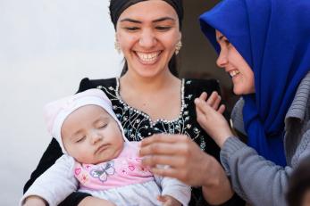 Two smiling women. The woman on the left is holding a sleeping baby.