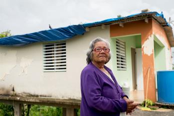 A woman with gray hair and a purple cardigan stands outside a home in Puerto Rico